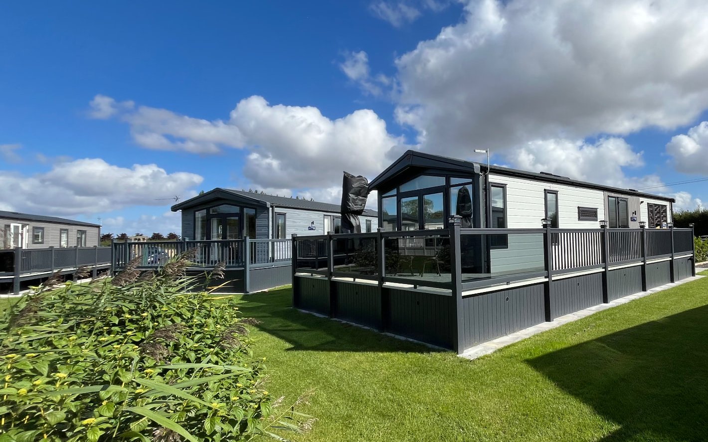 Lodges with dark grey glass-panelled decking on mown lawns at Croft Lakes Holiday Park under a bright blue sky with scattered cloud