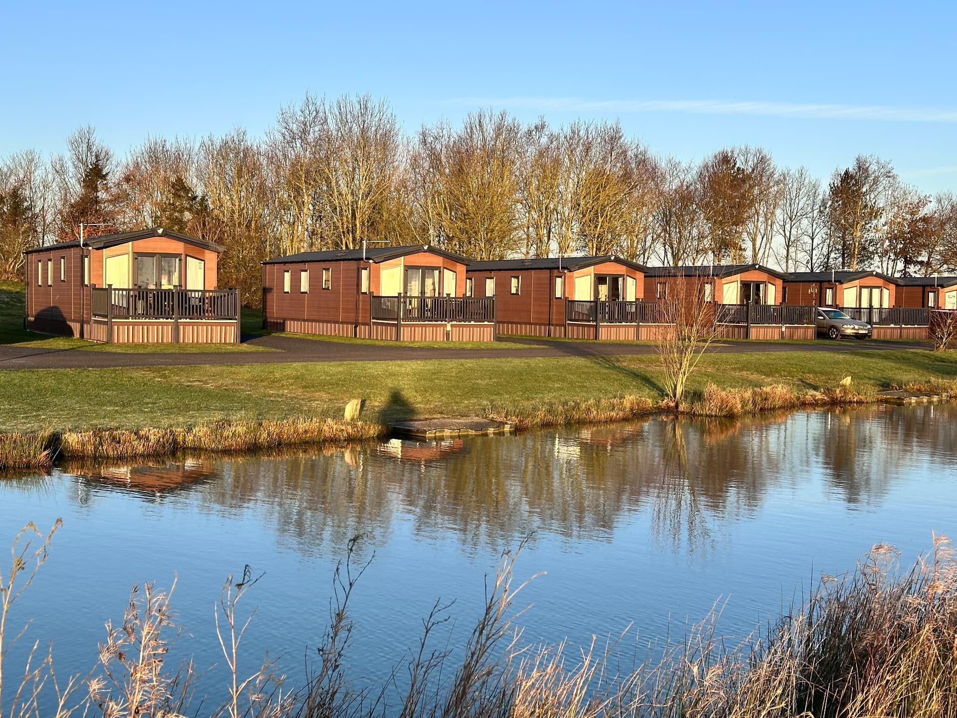 Row of timber-clad lodges and static caravans reflected in the lake at Hanworth Country Park in low winter sunshine, with bare trees behind