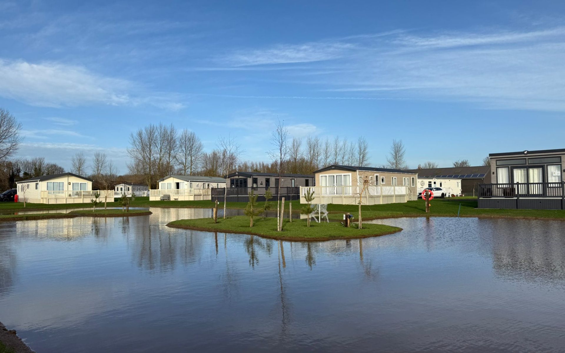 View across the lake at Virley House Country Park towards a row of static caravans and lodges, with a planted island in the water and clear blue sky above
